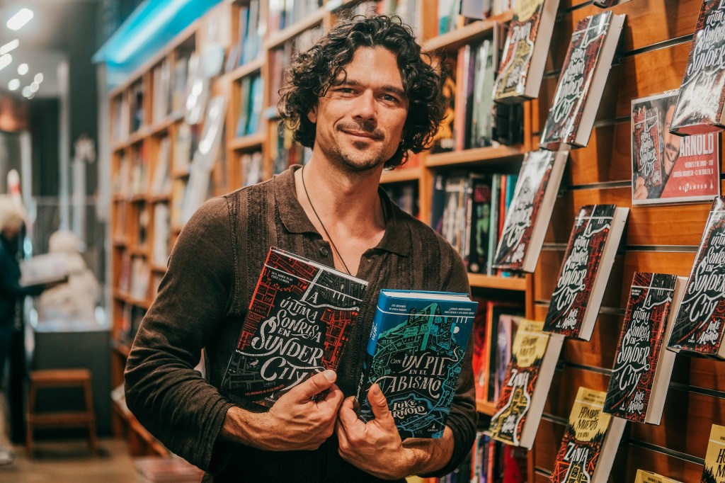 Fotografía de Luke Arnold posando junto con La última sonrisa en Sunder City y Con un pie en el abismo en la librería Gigamesh