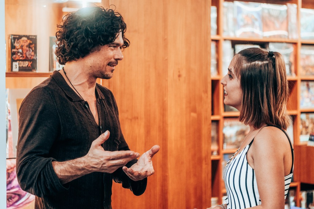 Fotografía de perfil en la que Luke Arnold y yo estamos hablando, de pie, en la librería Gigamesh. Luke está gesticulando con las manos.